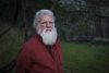 A photo of Bruce Pascoe. He stands outside and is wearing a red shirt.
