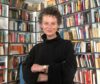 An author photograph of Andrea Goldsmith, who is standing in front of a bookshelf