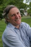 an author headshot of John Marsden. He is wearing a blue shirt and is pictured from his chest up. He is in 3/4 view, looking directly at the camera and smiling broadly. The photo was taken outside, and there is an out-of-focus lawn behind him. John is a white man with brown and grey hair and light-coloured eyes.