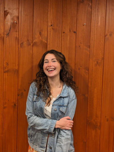 Ashleigh Mounser stands before a wooden wall with one arm crossed. She is wearing a denim jacket and smiling at the camera.