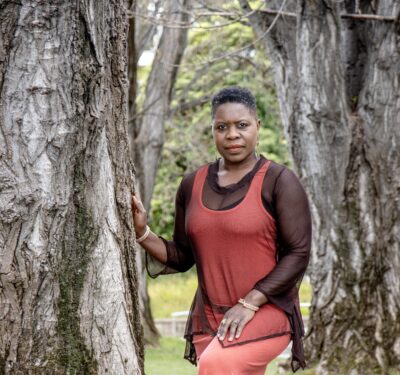 A portrait of Eugen Bacon, an African Australian woman wearing a red dress and standing between two trees