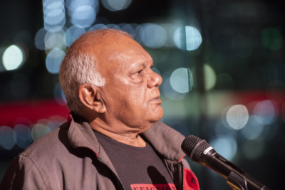 A photo of Lionel Fogarty taken by Leif Ekstrom. Fogarty stands before a microphone.