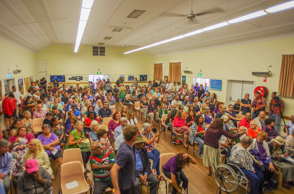 A community hall is filled with people seated, surrounded by others standing. Many are talking to one another or otherwise interacting; the image appears to have been taken before or after the event.